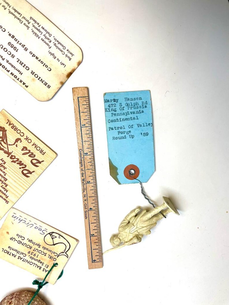 May include: A collection of vintage Girl Scout memorabilia, including tags, a wooden ruler, and a small plastic toy soldier. The tags feature handwritten text and the words "Sea Urchin" and "Round Up". The ruler measures up to 6 inches.