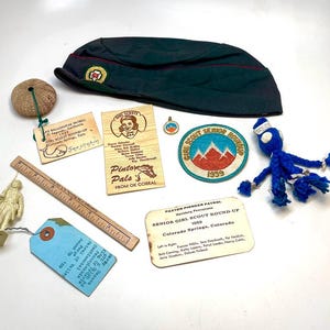 May include: A collection of vintage Girl Scout memorabilia, including a dark green cap, patches, wooden tags, a ruler, and a blue yarn doll. The items are arranged on a white surface, showcasing a nostalgic theme.