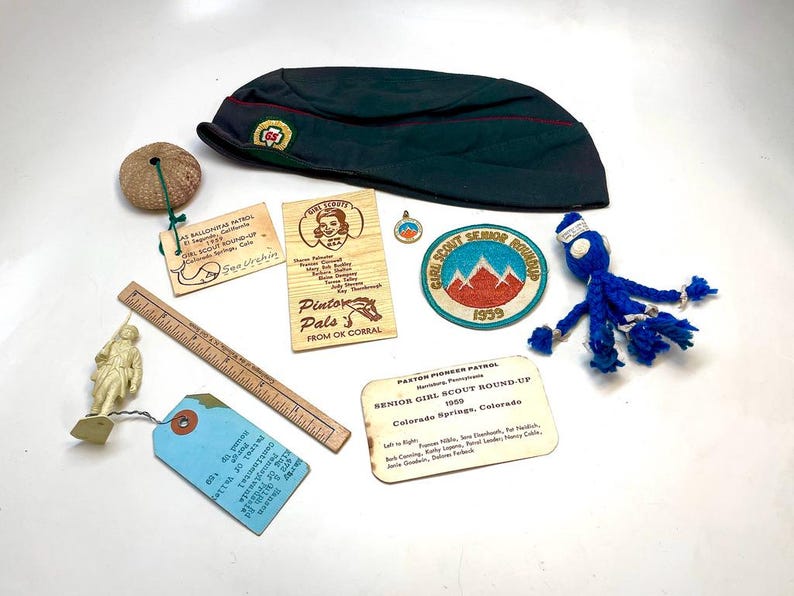 May include: A collection of vintage Girl Scout memorabilia, including a dark green cap, patches, wooden tags, a ruler, and a blue yarn doll. The items are arranged on a white surface, showcasing a nostalgic theme.