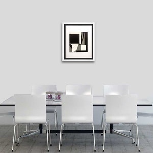 May include: A framed abstract art print with a black and white geometric design hangs above a white dining table. The artwork features angular shapes and contrasting tones. Six white chairs with metal legs are positioned around the table, creating a modern dining room setting.