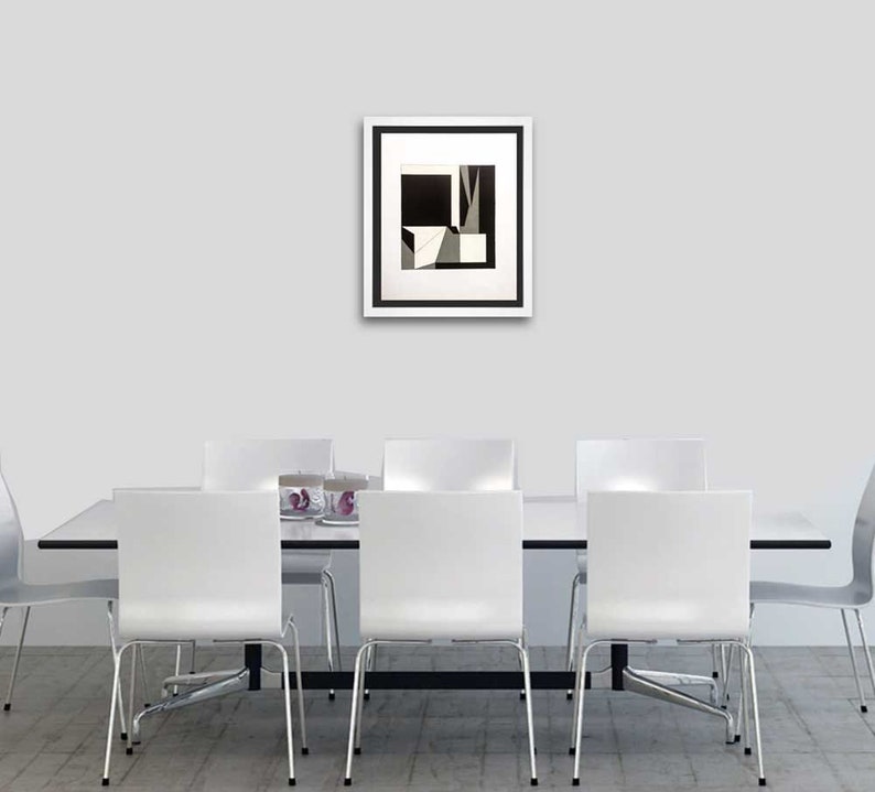 May include: A framed abstract art print with a black and white geometric design hangs above a white dining table. The artwork features angular shapes and contrasting tones. Six white chairs with metal legs are positioned around the table, creating a modern dining room setting.