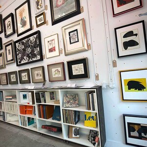 Puede incluir: Una estanter&iacute;a blanca con varios libros y obras de arte enmarcadas en la pared detr&aacute;s de ella. Las obras de arte son una mezcla de impresiones en blanco y negro y en color.
