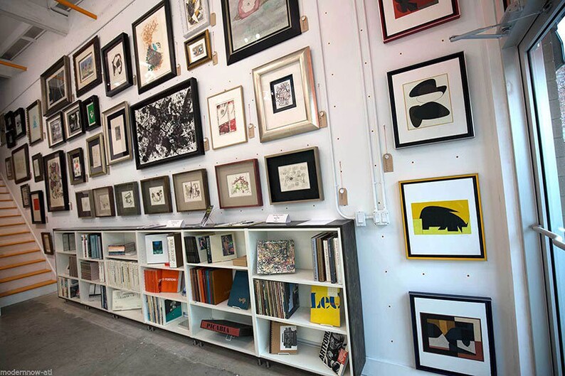 Puede incluir: Una estanter&iacute;a blanca con varios libros y obras de arte enmarcadas en la pared detr&aacute;s de ella. Las obras de arte son una mezcla de impresiones en blanco y negro y en color.