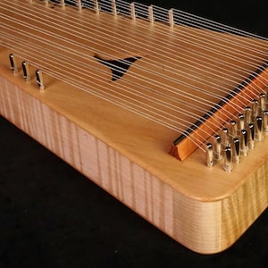 Könnte beinhalten: Ein helles, hölzernes Saiteninstrument im Zither-Stil mit einer glatten, polierten Oberfläche und sichtbarer Holzmaserung. Es besitzt zahlreiche dünne Metallsaiten, die über den Korpus gespannt und durch silberne Stimmwirbel sowie einen Holzsteg befestigt sind.