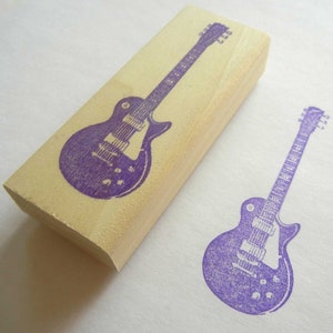 May include: A wooden block stamp with a purple electric guitar design. The stamp is used to create a print of the guitar on paper.