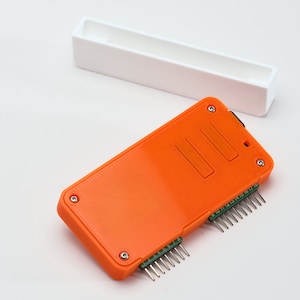 以下が含まれることがあります： 白のプラスチック製の蓋が付いたオレンジ色のプラスチック製のケース。ケースの片側にはピンが複数あり、上部と下部には4本のネジがあります。