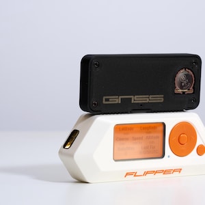 May include: Two-part electronic device. The top section is black with "GNSS" printed on it. The bottom section is white with an orange screen and button. The word "FLIPPER" is printed on the bottom.