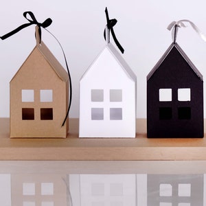 May include: Three paper houses with cut-out windows, each in a different color: brown, white, and black. Each house has a black ribbon tied around the top.