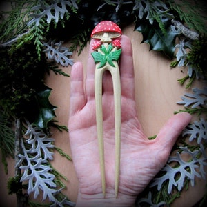 May include: A wooden hair stick with a carved mushroom design. The mushroom is red with white spots and green leaves. The stick is light brown and has a smooth finish.