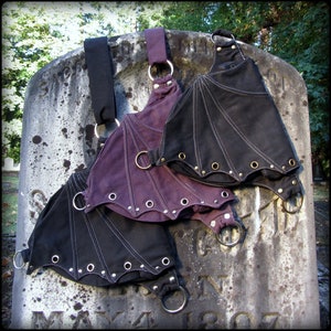 May include: Three black and purple bat-shaped bags with straps and metal rings. The bags are made of canvas and have a gothic style.