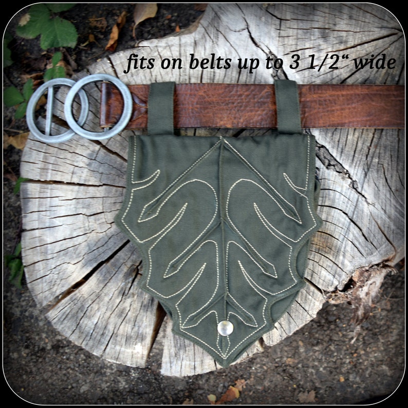 May include: A green leaf-shaped pouch with a button closure. The pouch is made of fabric and has a stitched design. It is attached to a brown leather belt with the text "fits on belts up to 3 1/2" wide" printed on the belt.