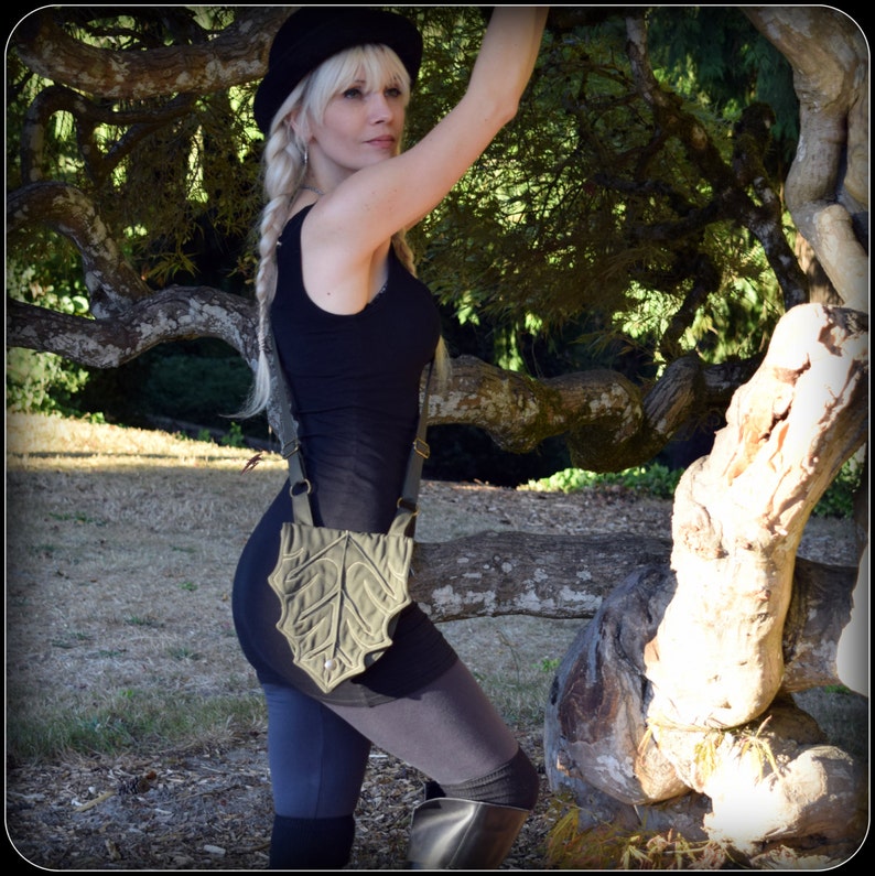 May include: A woman wearing a black tank top and black pants with a green leaf-shaped crossbody bag. She is standing in front of a tree with a large trunk.