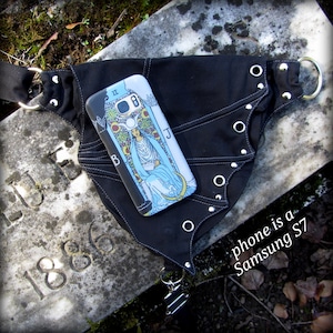 May include: A black fabric belt with silver grommets and a phone case featuring the High Priestess card from a tarot deck. The phone is a Samsung S7.