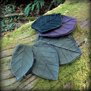 May include: Four leaf-shaped fabric pouches in different sizes. The largest pouch is green, the next is purple, and the smallest two are dark green. The pouches are arranged in a stack on a mossy log.