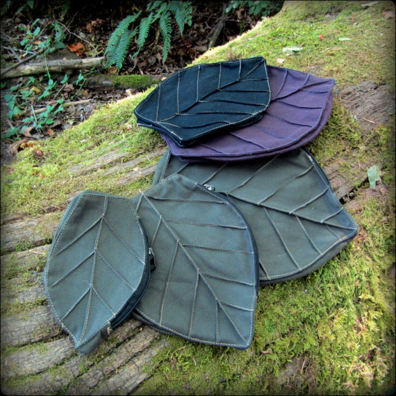 May include: Four leaf-shaped fabric pouches in different sizes. The largest pouch is green, the next is purple, and the smallest two are dark green. The pouches are arranged in a stack on a mossy log.