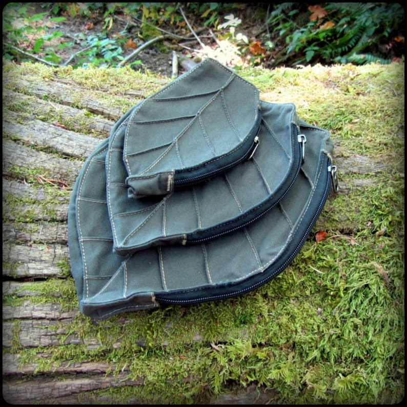 May include: Three gray canvas leaf-shaped pouches with zipper closures, stacked on top of each other on a mossy log.