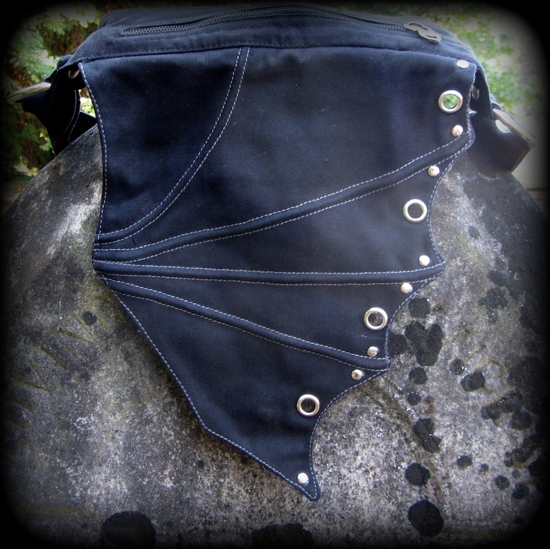 May include: Black canvas bag with a wing-shaped flap. The flap has white stitching and silver grommets and studs.