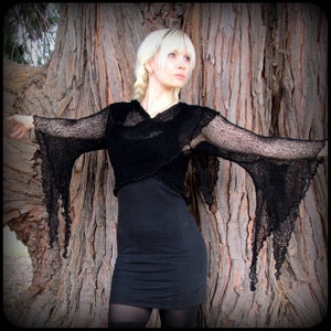 May include: A black sheer top with long, flowing sleeves and a draped neckline. The top is worn over a black dress and the model is standing in front of a tree.