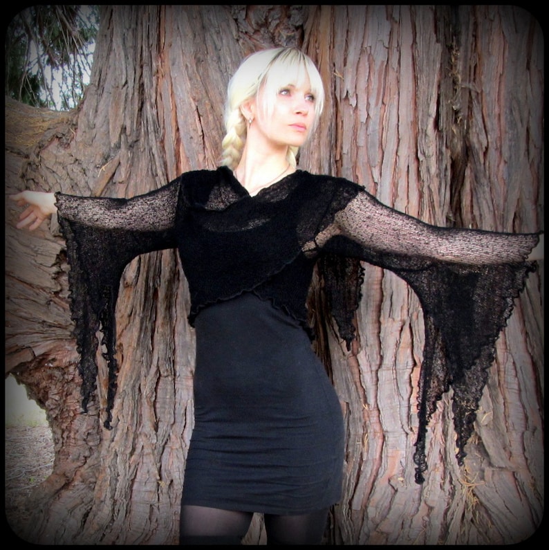 May include: A black sheer top with long, flowing sleeves and a draped neckline. The top is worn over a black dress and the model is standing in front of a tree.