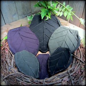 May include: Six leaf-shaped zippered pouches in various shades of green, black, and purple. The pouches are arranged in a nest of twigs.