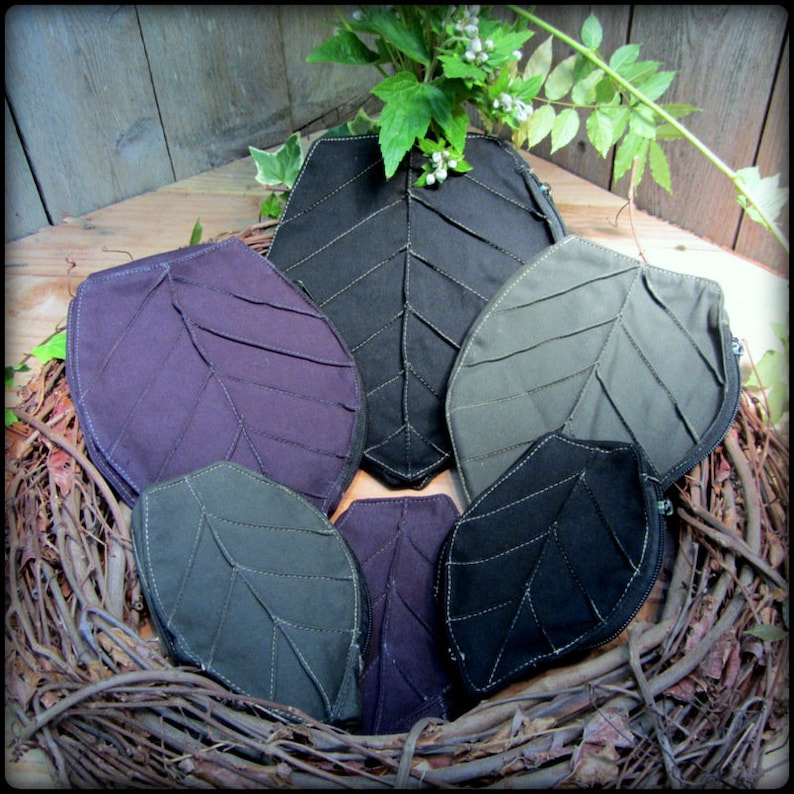 May include: Six leaf-shaped zippered pouches in various shades of green, black, and purple. The pouches are arranged in a nest of twigs.