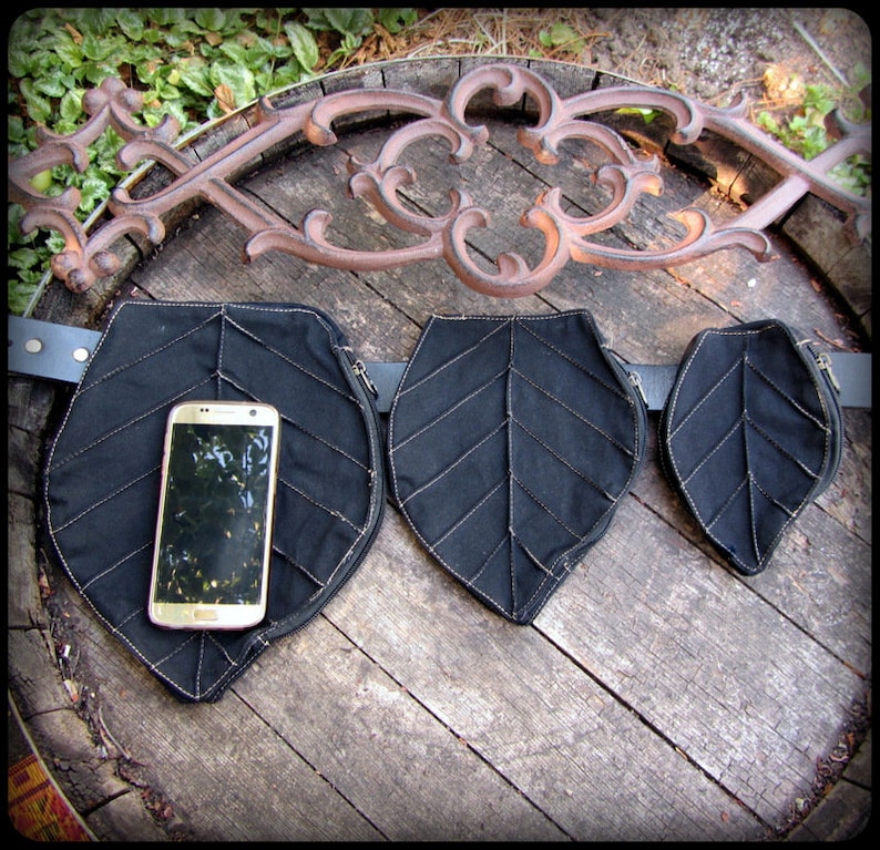 May include: A black leather belt with three leaf-shaped pouches. The pouches have zippers and are lined with black fabric. A gold smartphone is in the left pouch.