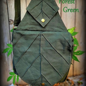 May include: A forest green canvas leaf-shaped crossbody bag with a flap closure and two gold-toned metal buttons. The bag has a strap for carrying over the shoulder.