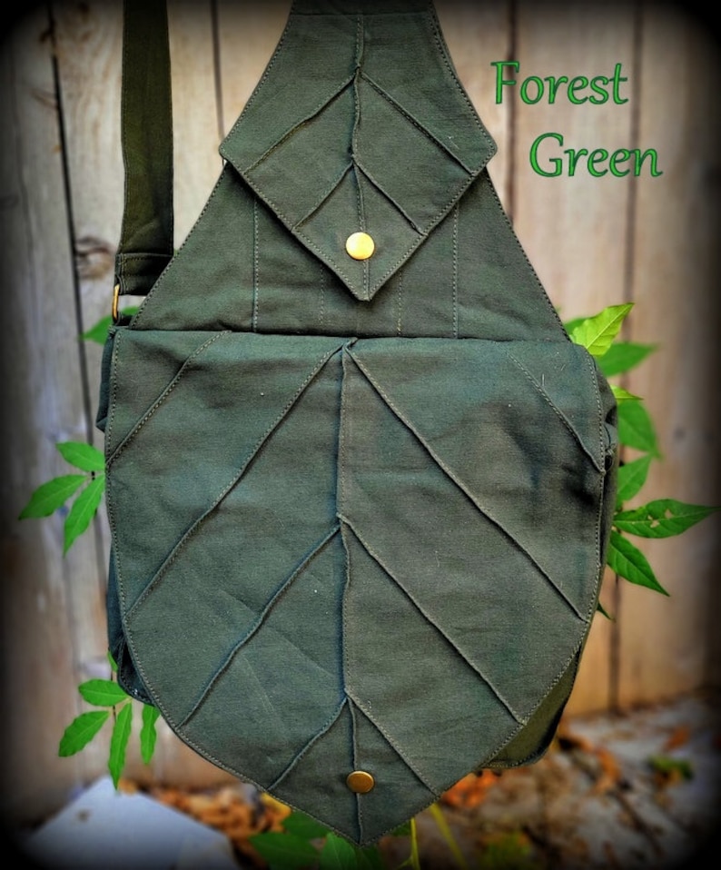 May include: A forest green canvas leaf-shaped crossbody bag with a flap closure and two gold-toned metal buttons. The bag has a strap for carrying over the shoulder.