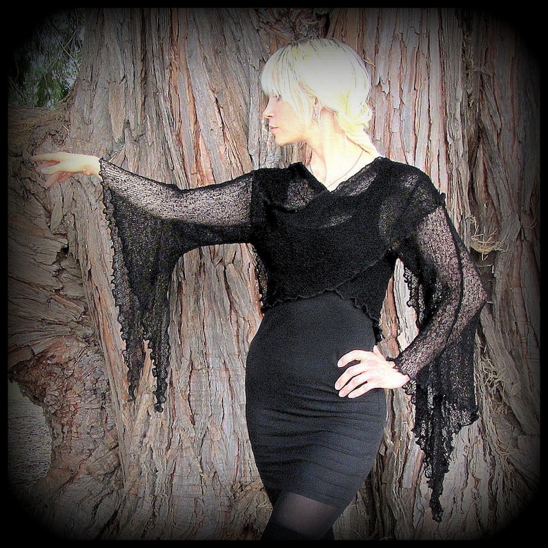 May include: A black sheer top with long, flowing sleeves and a draped neckline. The top is worn over a black dress and the model is standing in front of a tree.