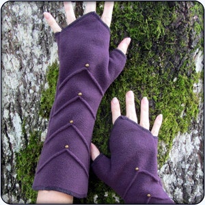 Könnte beinhalten: Lilafarbene Fingerlose Fleecehandschuhe mit dekorativen Nähten und goldfarbenen Akzenten. Die Handschuhe haben eine lange Manschette und sind so konzipiert, dass sie Handgelenk und Unterarm bedecken.