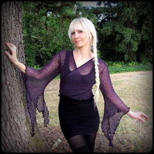 May include: A woman wearing a purple, sheer, long-sleeved top with a ruffled hem and a black skirt. She has long blonde hair braided into two braids.