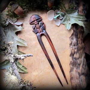 May include: A wooden hair fork with a carved mushroom design. The fork is dark brown with a reddish-brown mushroom and leaves.