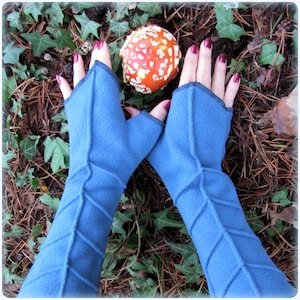 Könnte beinhalten: Ein Paar blaue Fleece-Fingerhandschuhe mit einem strukturierten Design. Die Handschuhe werden von einer Person mit rotem Nagellack getragen. Die Handschuhe befinden sich auf einem Hintergrund aus braunen Kiefernnadeln und grünen Blättern.