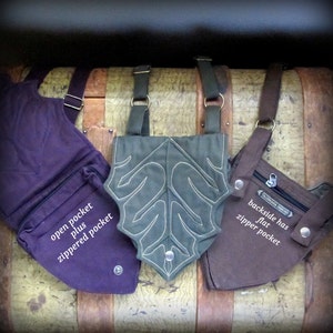 May include: Three different canvas bags with straps. The bag on the left is purple with an open pocket and a zippered pocket. The bag in the middle is green with a leaf design and a zippered pocket. The bag on the right is brown with a zippered pocket and the text "backside has flat zipper pocket".