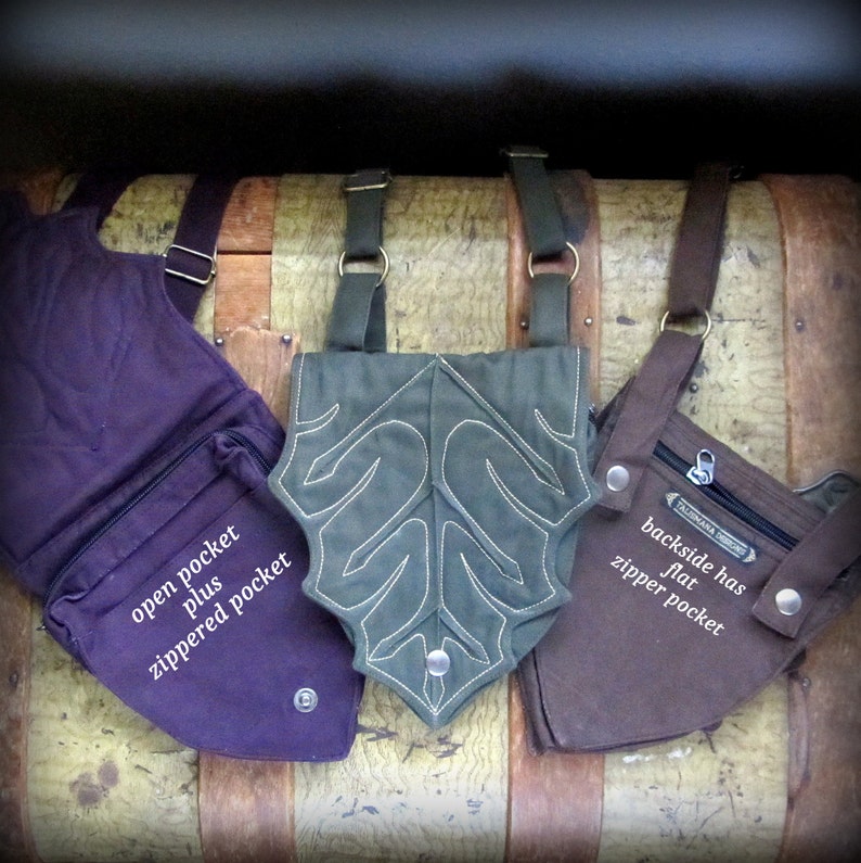 May include: Three different canvas bags with straps. The bag on the left is purple with an open pocket and a zippered pocket. The bag in the middle is green with a leaf design and a zippered pocket. The bag on the right is brown with a zippered pocket and the text "backside has flat zipper pocket".