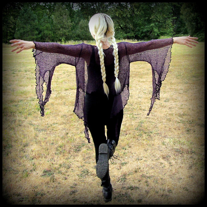 May include: A woman with long blonde braids is wearing a long, dark purple, sheer, open-weave top with long sleeves. She is standing in a field with her arms outstretched.