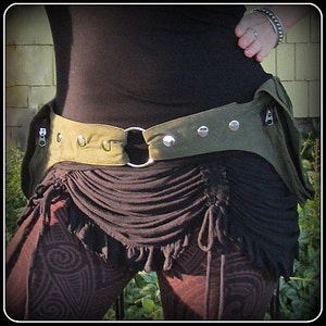 Op de afbeelding: Een groene canvas riem met metalen accenten en een ritsvak. De riem wordt gedragen over een zwart topje en een bruine rok met een gerimpelde zoom.