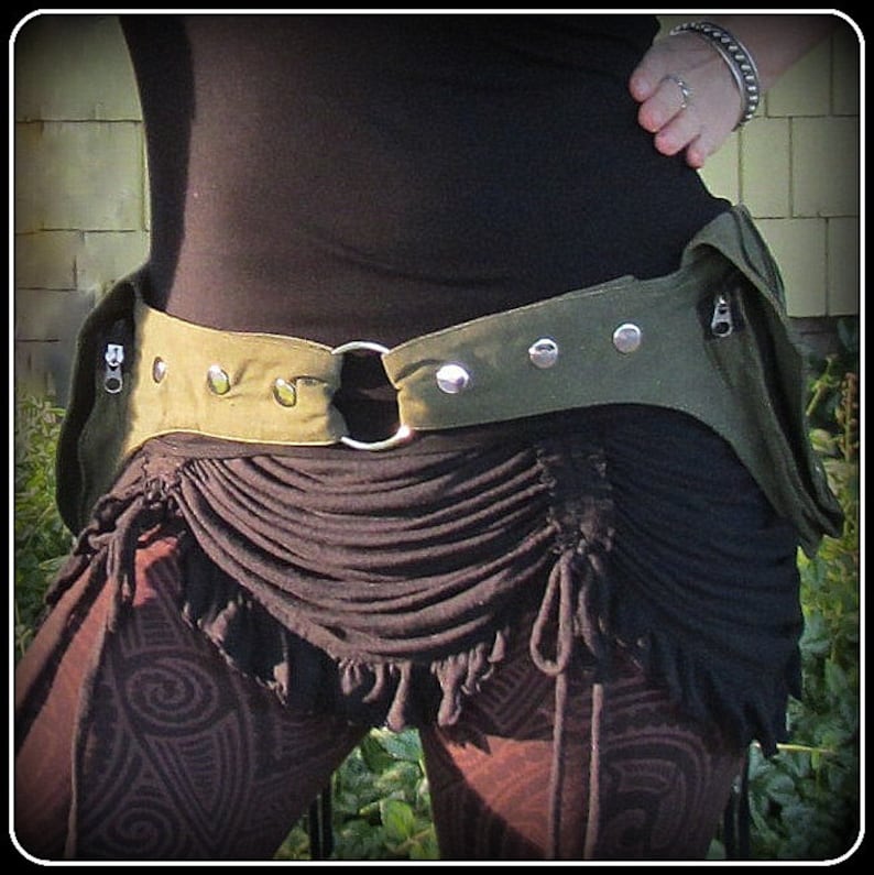 Op de afbeelding: Een groene canvas riem met metalen accenten en een ritsvak. De riem wordt gedragen over een zwart topje en een bruine rok met een gerimpelde zoom.