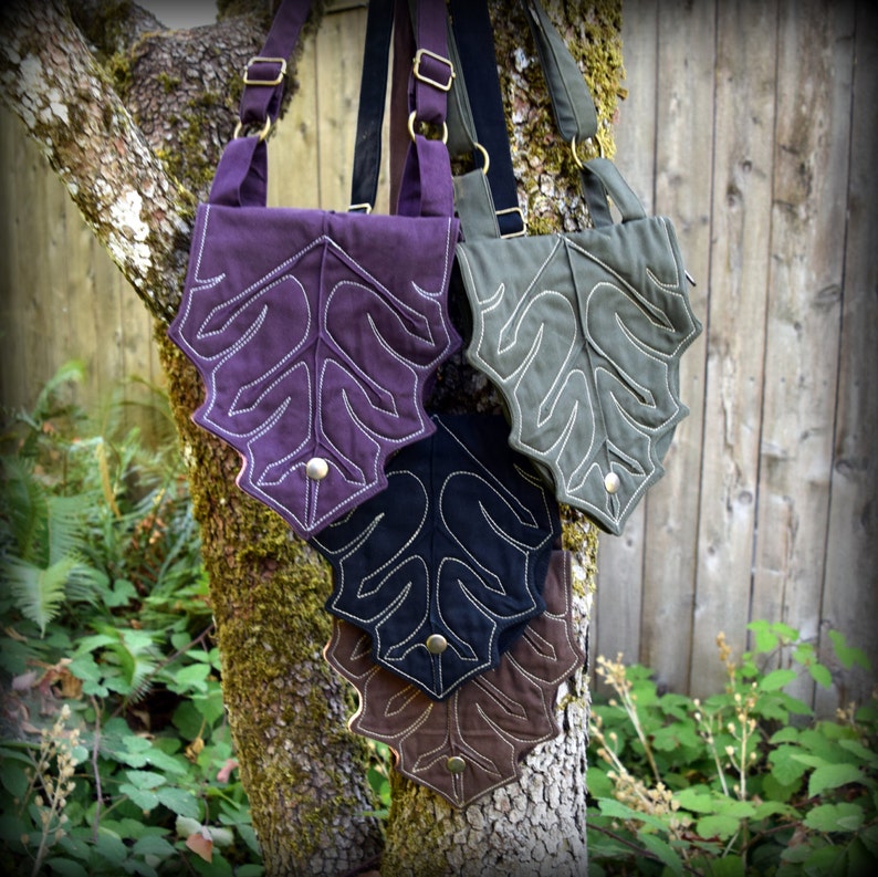 May include: Four leaf-shaped crossbody bags in different colors: purple, black, green, and brown. Each bag has a white stitched leaf design and a metal button.