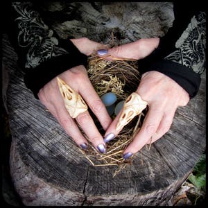 May include: Two bone rings shaped like bird skulls with intricate details. The rings are worn on the index and middle fingers of a person's hand. The hand is resting on a nest of twigs and straw.