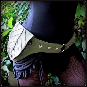 Op de afbeelding: Een groene canvas riem met een blaadjesvormige tas en zilveren metalen accenten. De riem wordt om de taille gedragen en heeft een rits op de tas.