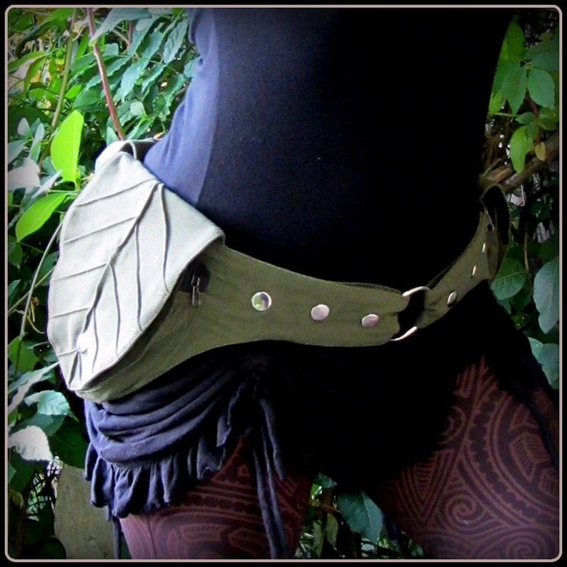 Op de afbeelding: Een groene canvas riem met een blaadjesvormige tas en zilveren metalen accenten. De riem wordt om de taille gedragen en heeft een rits op de tas.