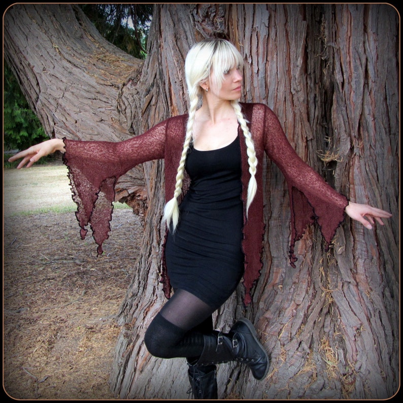 May include: A woman with long blonde braids wears a maroon lace cardigan with long, flowing sleeves. She is standing in front of a tree trunk and wearing a black dress and black tights.