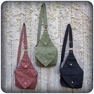 May include: Three canvas crossbody bags with leaf-shaped flap closures. The bags are in green, brown, and black.