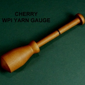 May include: A wooden yarn gauge with a cherry wood finish. The gauge has a black line marking the measurement. The text "CHERRY WPI YARN GAUGE" is printed on a green background.