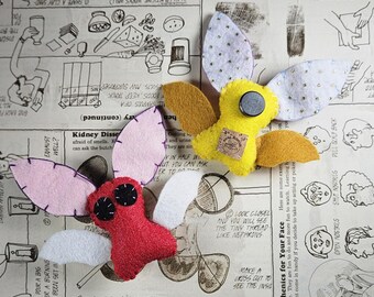 May include: Two plush insect-shaped ornaments. One is red with black stitched eyes and light pink and white wings. The other is yellow with gold wings, white dotted wings, a black circular center, and a small square tag labeled Scrap Happy.