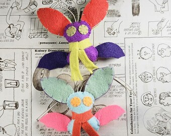 May include: Two handmade felt moth plushies with stitched details. The top moth has red wings, a purple body, yellow scarf, and beige eyes. The bottom moth has light green wings, a light blue body, orange scarf, and orange eyes.