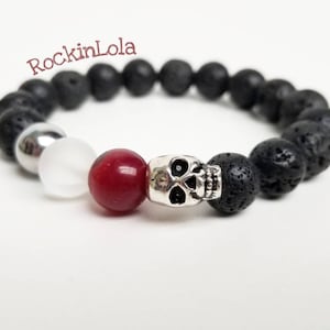 May include: A black lava stone bead bracelet with a silver skull charm, a red bead, a white bead, and a silver bead. The bracelet has the text "RockinLola" in the upper left corner.