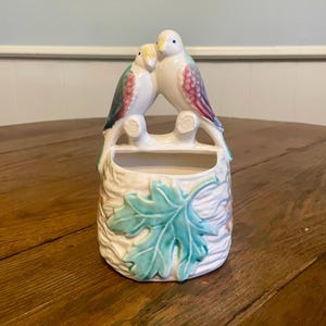May include: A ceramic wall pocket featuring two perched birds with white, teal, and purple accents. The birds sit atop a textured, off-white base with a teal leaf design. The pocket is designed for small plants or decorative items.