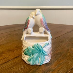 May include: A white ceramic wall pocket featuring two perched birds with colorful wings. The birds are atop a small, rectangular opening. The base of the pocket has a leaf design with green and brown accents. The piece is on a wooden surface.